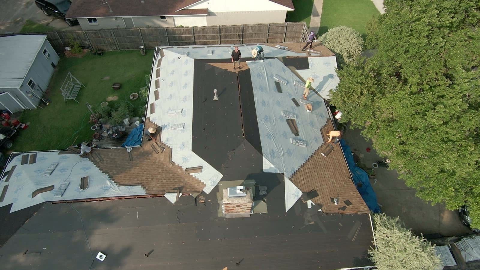 Overhead drone view of a roofing crew rolling out synthetic underlayment while old brown shingles are torn off the rest of the roof