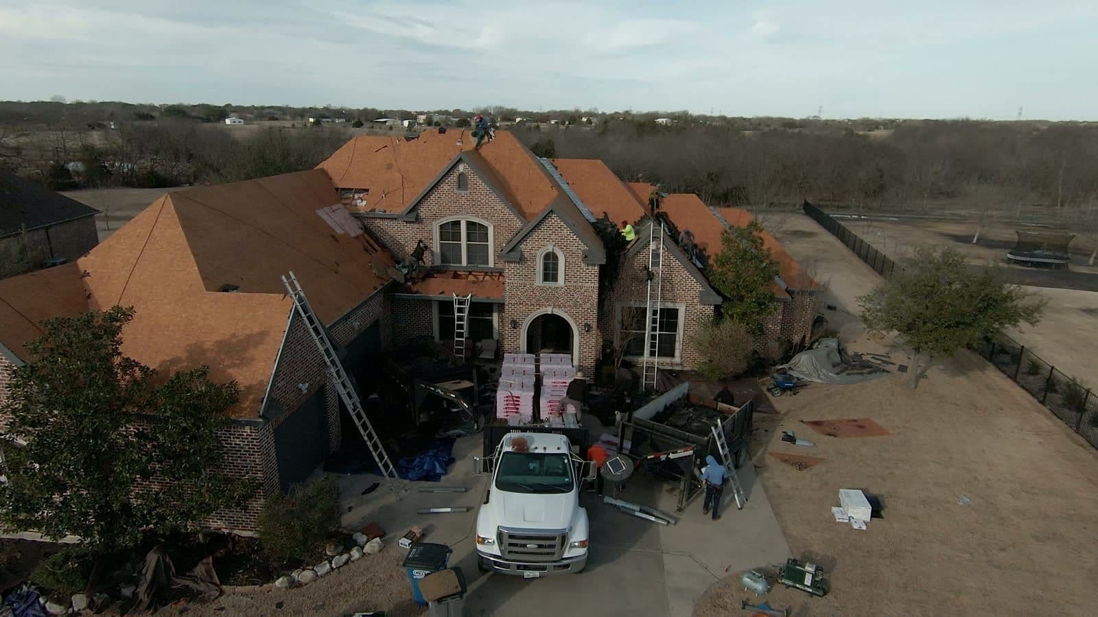 Large brick two-story home mid tear-off with the roof decking exposed, ladders against the walls and shingle bundles in the driveway