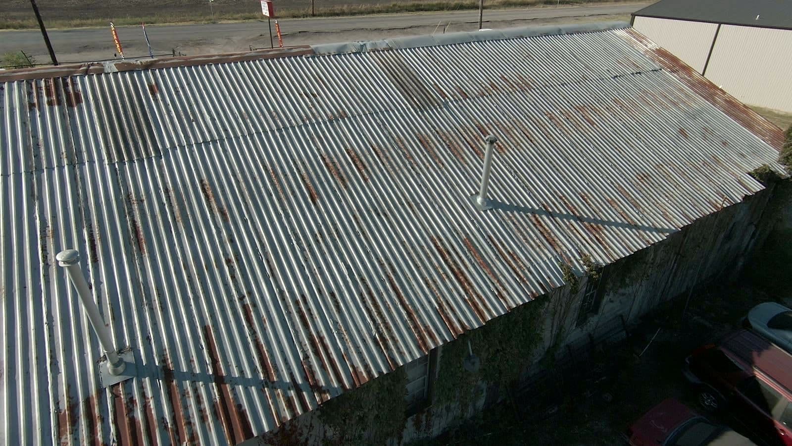 Rusted corrugated metal roof on a commercial building in Royse City, with vent pipes and a road beyond