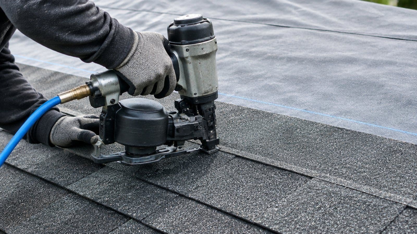 Close-up of a roofer's gloved hands nailing a new charcoal architectural shingle into place over synthetic underlayment
