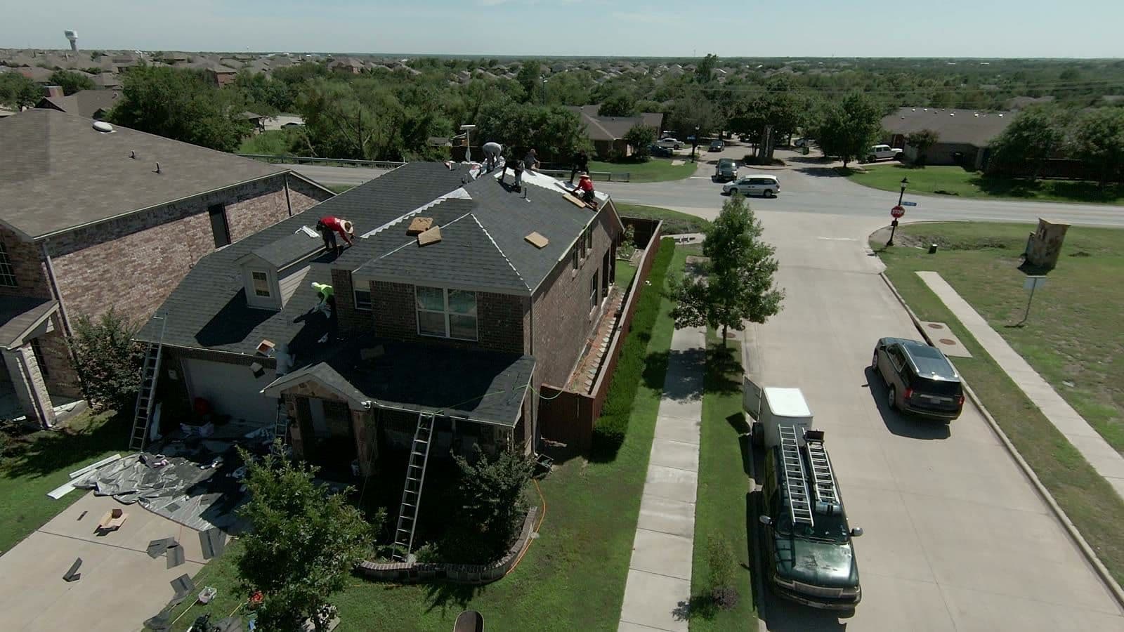 Roofing crew replacing shingles on a brick two-story home on a neighborhood corner, with ladders against the walls